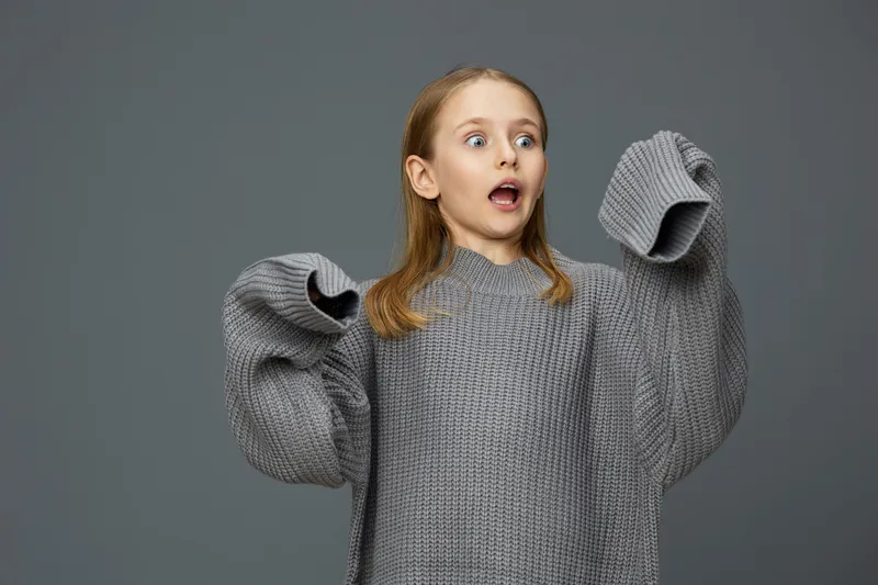 Surprised young girl in oversized grey sweater, expressing excitement against a muted background, capturing emotions of joy and surprise, perfect for seasonal campaigns