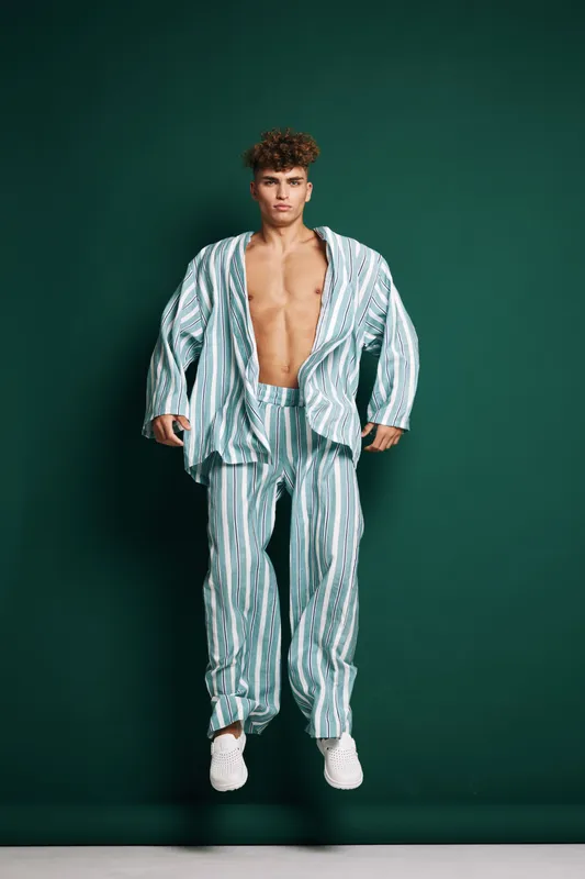 Young man with curly hair wearing striped pajama set and white sneakers standing against dark green background with open shirt showing bare chest and confident pose.