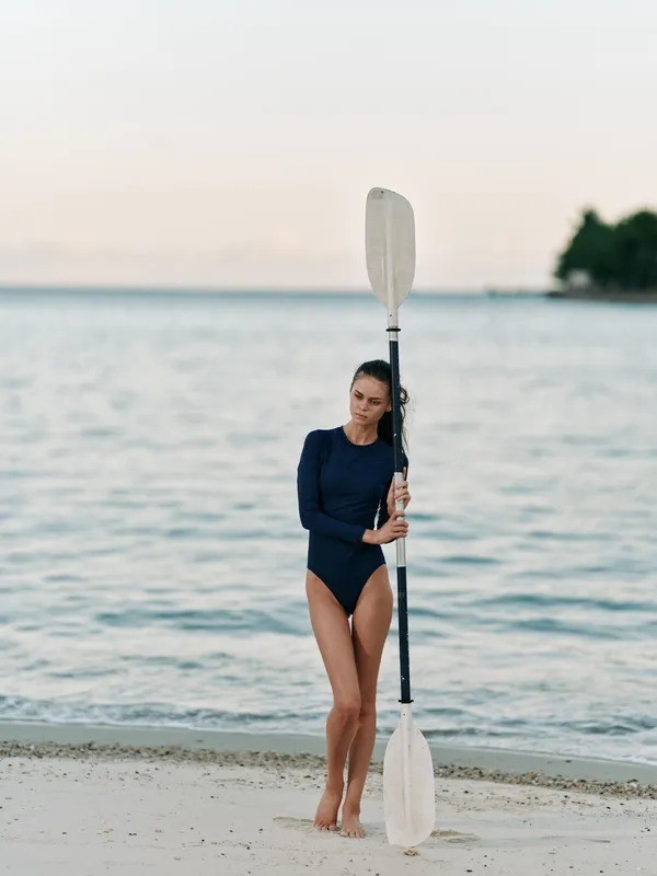 Female athlete in a navy swimsuit stands on a sandy beach, gripping a double paddle upright as a calm sea and pastel sky set a tranquil scene.