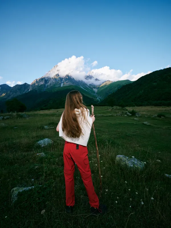 Woman in red pants standing in a wide grassy meadow with a walking stick, facing distant mountains under a bright blue sky, capturing expansive nature, open landscape, and outdoor adventure vibes
