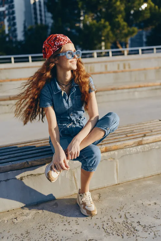 Lifestyle woman in modern boho-western denim street style with flowing curly hair, red bandana, and sunglasses, captured in warm film color under natural sunlight.
