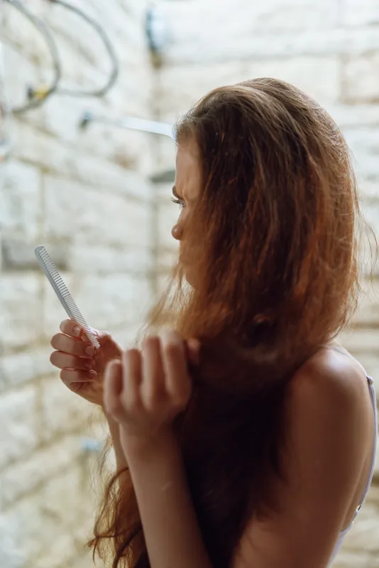 woman with long hair combing wet strands in a bright bathroom with beige tiles, natural light illuminating healthy hair strands during morning routine