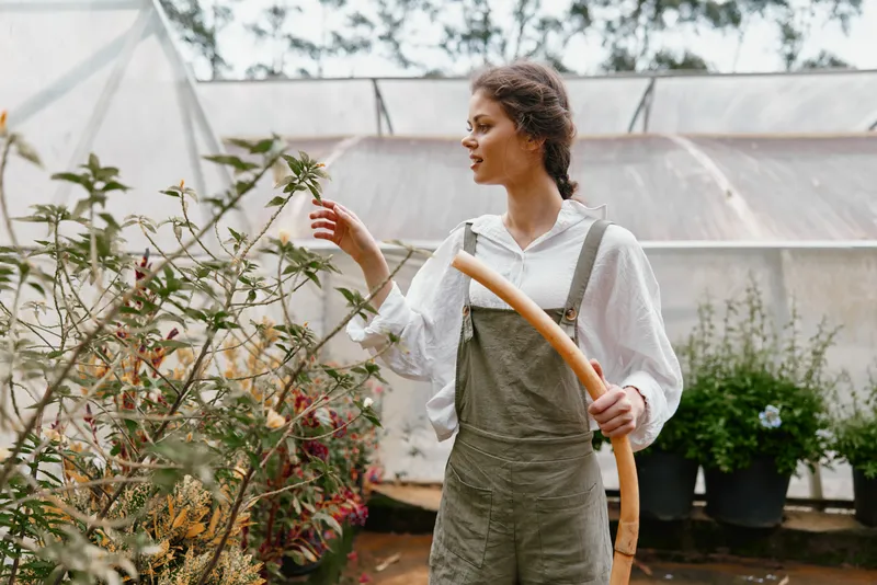 Young woman in greenhouse, wearing light overalls and a white shirt, showing joy while gardening, surrounded by lush plants, autumn colors, and natural light
