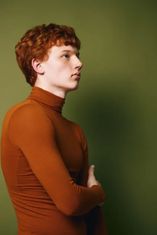 Young man with red hair wearing brown turtleneck sweater posing in profile on green background. Casual style, thoughtful expression, isolated studio portrait with natural light and soft shadows.