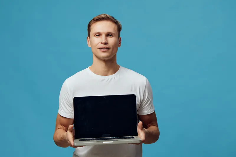 Smiling man holding open laptop computer in front of plain blue background studio portrait, casual clothing, young adult, positive expression, technology, modern work environment