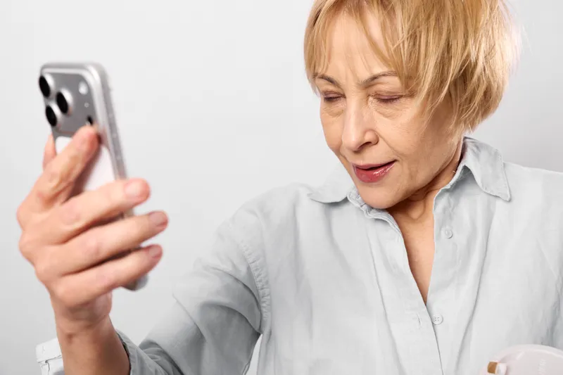 Elderly woman holds smartphone, surprised reading a message on white background, home communication, technology portrait, closeup view, soft studio light