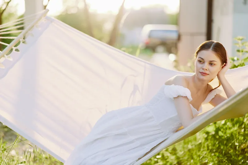 Young woman in a white summer dress relaxing on a hammock at sunset, surrounded by greenery with a soft, warm glow enhancing her peaceful expression
