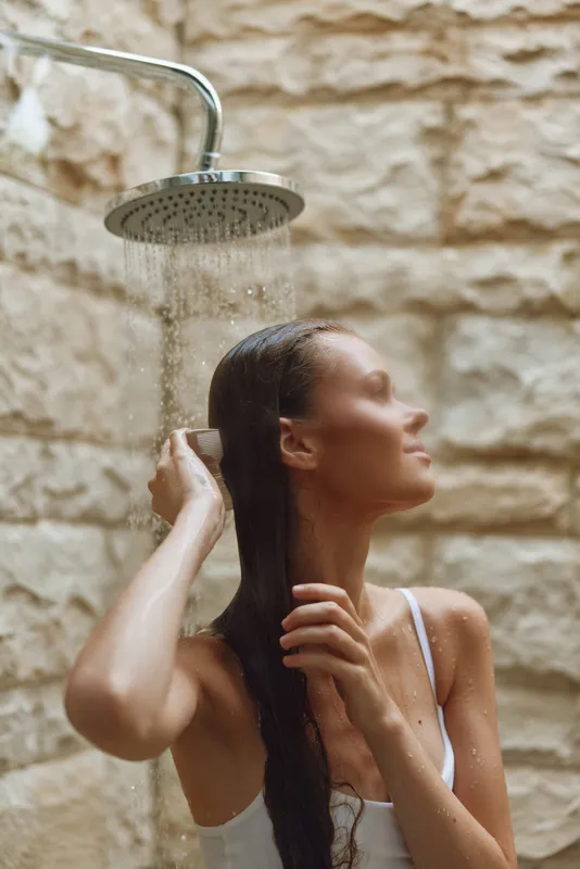 woman showering outdoors with wet hair near stone wall, natural light, serene expression, fresh water droplets, summer bathing