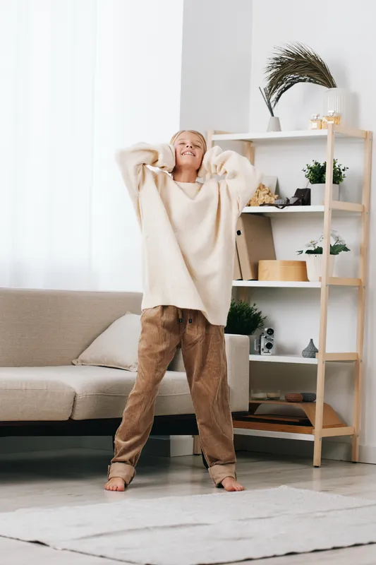 An authentic candid moment of home play, featuring a young student in cozy attire, stretching with joy in a bright living space that hints at daily school life.
