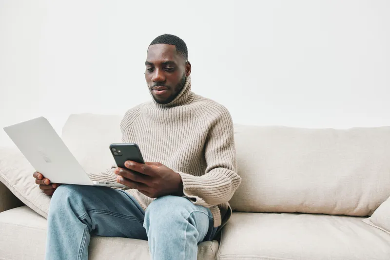 Young Black man in a stylish sweater using a laptop and smartphone on a modern couch, showcasing technology and communication, perfect for lifestyle concepts