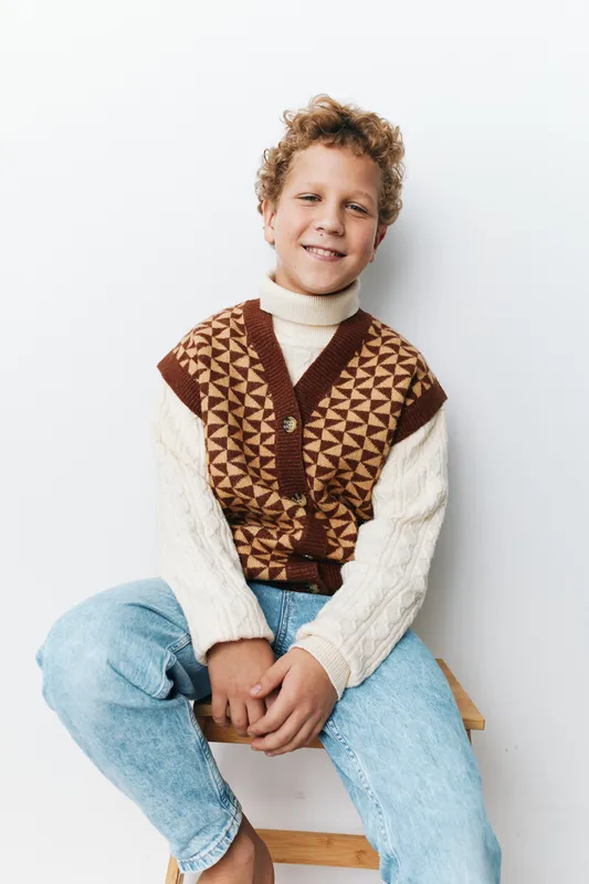 Portrait of a young boy seated on a wooden stool, wearing a patterned brown knit vest over a cream turtleneck and light jeans, smiling softly against a white backdrop.