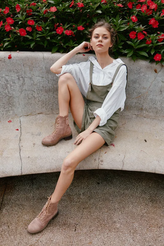 A stylish young model poses gracefully in a garden, wearing khaki overalls and a white blouse, exuding confidence amidst vibrant flowers