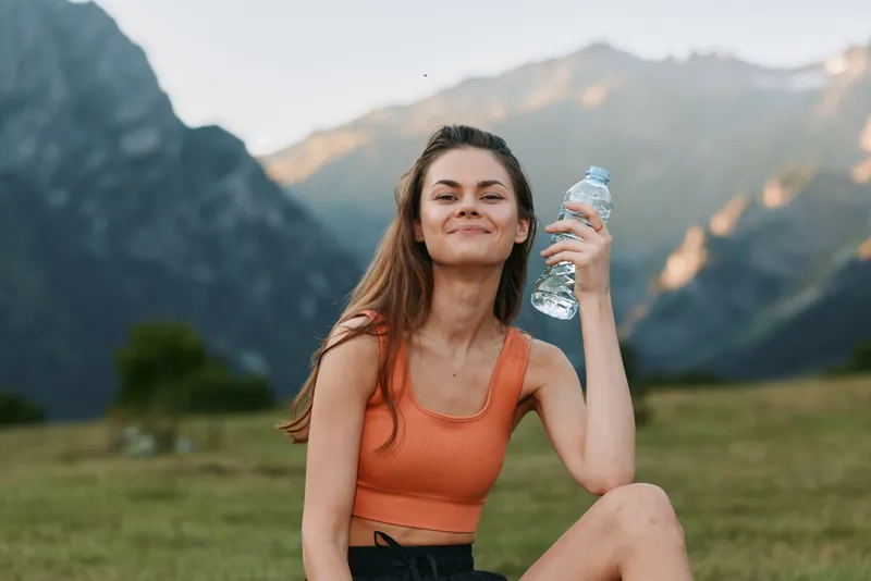 A cheerful outdoorswoman rests on grass in a mountain landscape. She wears an orange sports top, smiles confidently, and holds a water bottle, exuding fresh energy and wellness.