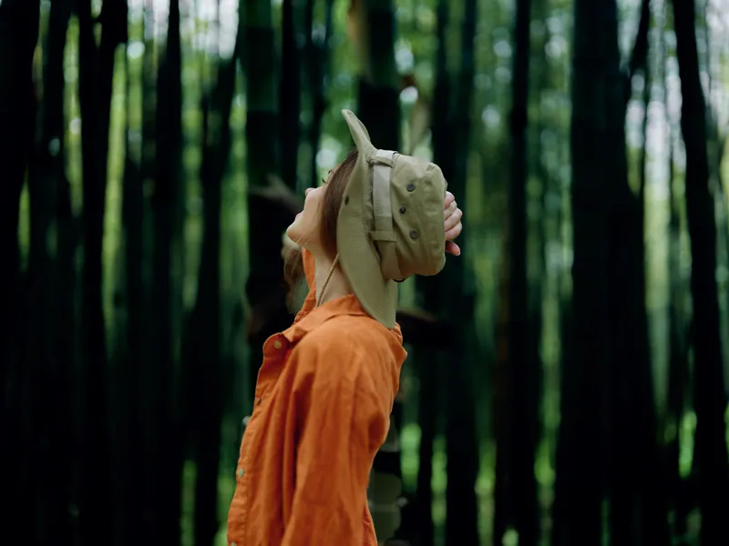 Forest woman in orange jacket and hat looks upward among tall trees, nature portrait in profile view exploring woodland light and greenery, outdoor travel mood and quiet solitude.