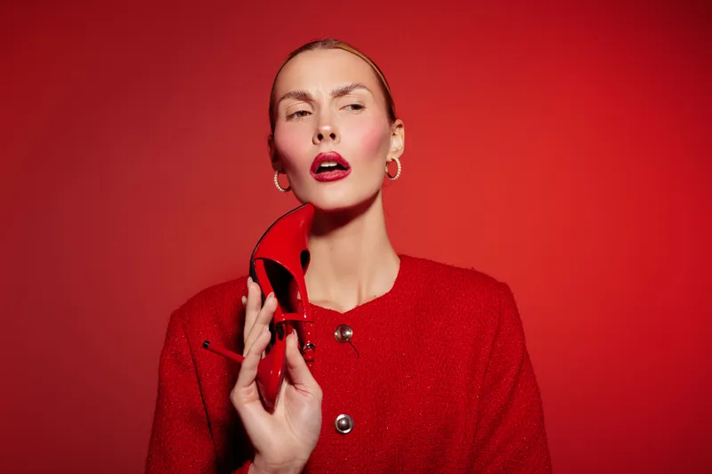 Stylish woman in red jacket holds matching high heel shoe near face against red background. Fashion concept with bold colors and confident expression in studio lighting.