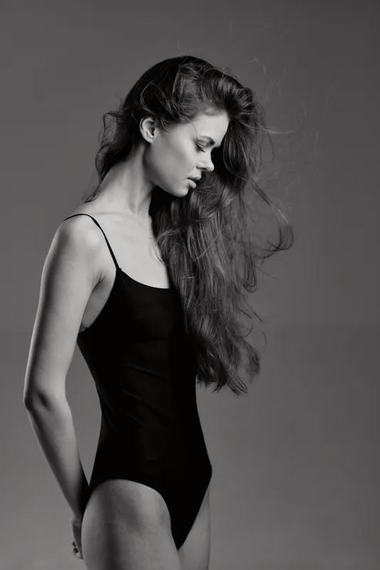 Black and white portrait of a thoughtful woman in a black leotard with long hair against plain background.
