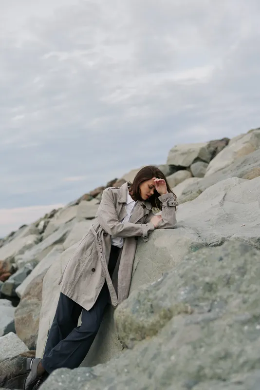 A thoughtful woman in a long coat leans on large rocks by the sea under a cloudy sky. The mood is calm and reflective, capturing solitude and nature connection.