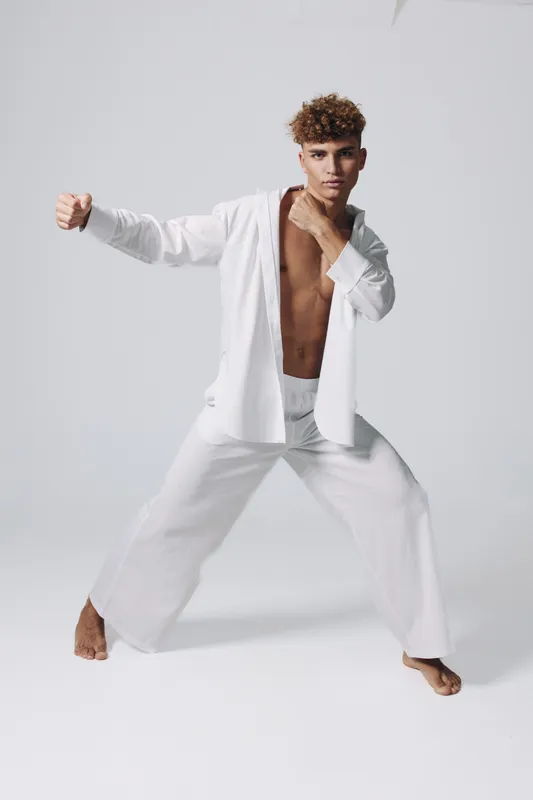 Young man practicing martial arts in white loose pants and open shirt, demonstrating fighting stance and focus in studio with plain background and barefoot pose.