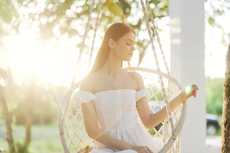 Young woman in a white dress sitting in a swing, enjoying a serene moment in nature during sunset, exuding tranquility and elegance Natural light enhances a romantic atmosphere