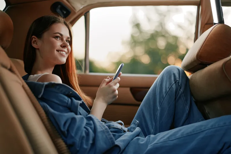 Young woman smiling while using a smartphone in a car, showcasing a relaxed and comfortable atmosphere with soft lighting and a casual outfit.