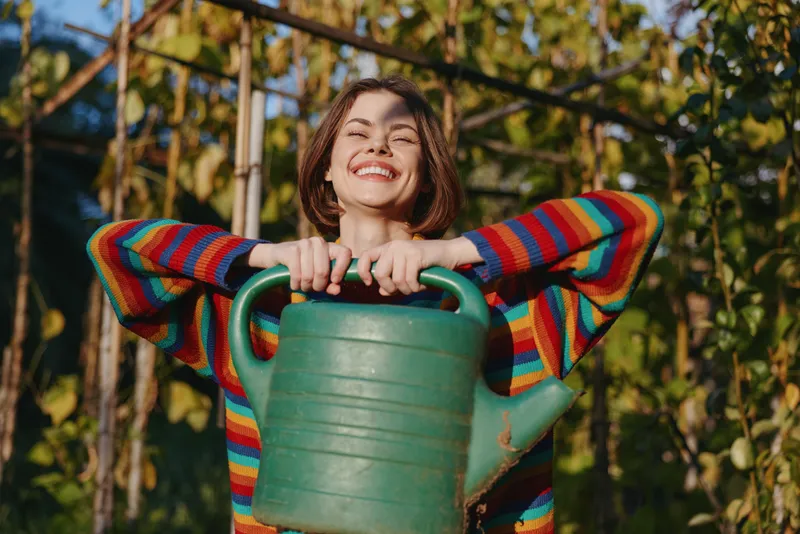 Woman holding a green watering can in garden, striped sweater and short brown hair, smiling joyfully. Young adult outdoor lifestyle, casual outfit, summer gardening and home care.