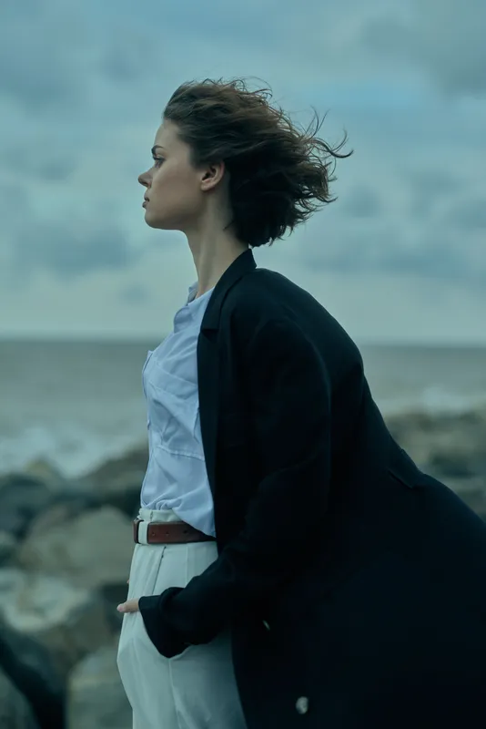 Wind, sea breeze, profile of a woman in a dark coat by the shoreline, windblown hair and calm ocean in the background, minimalist outdoor fashion mood, soft light, dramatic clouds