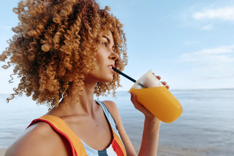 Smiling woman with curly hair sipping orange juice on a sunny beach, casual summer scene, refreshing drink, leisure mood, warm lighting and relaxed vibe.