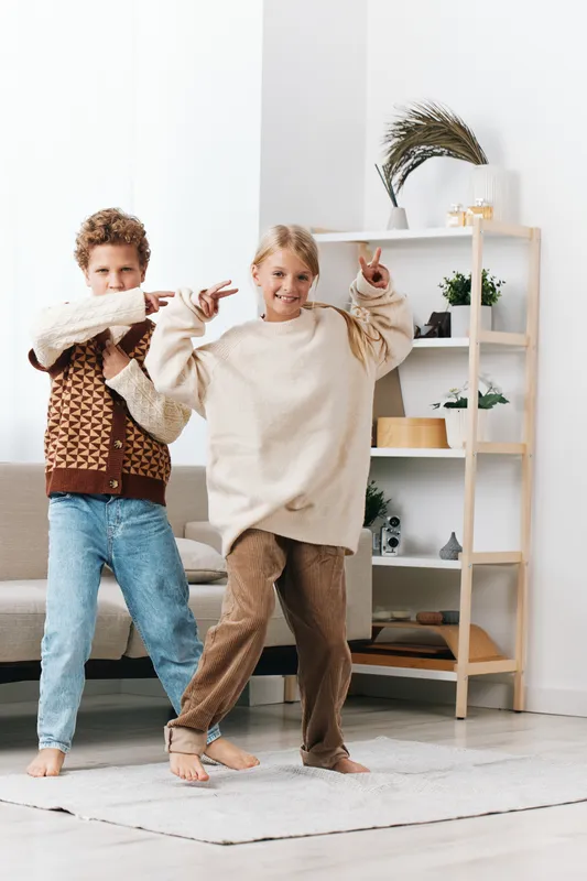 Two young children dance playfully at home, smiling and pointing, wearing cozy sweaters and jeans, in a bright room with soft natural light and minimal furniture, capturing joy.