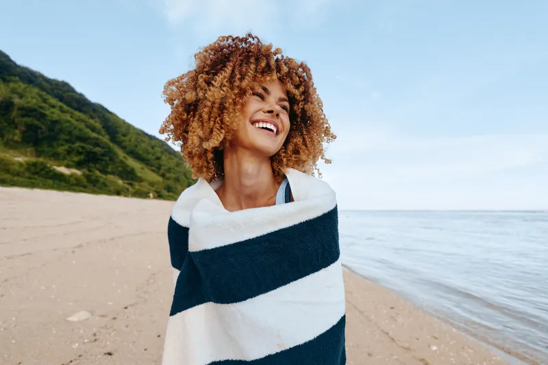 Young woman with curly hair wrapped in a striped towel smiles happily on a sandy beach near calm water under a clear sky.