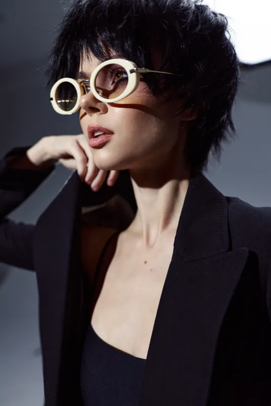 Stylish woman wearing round sunglasses and a black blazer in soft dramatic lighting.