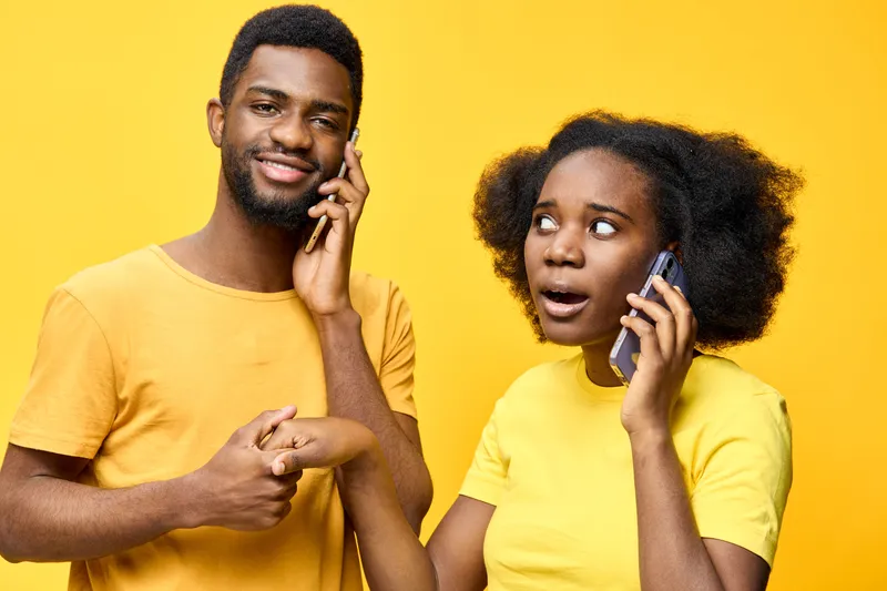 Cheerful young African couple talking on their smartphones against a vibrant yellow background, expressing surprise and joy, showcasing contemporary lifestyle