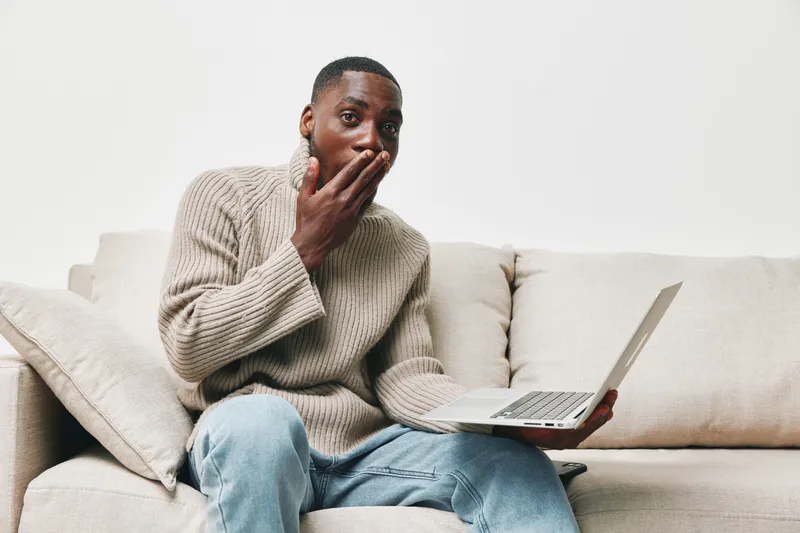 Surprised young African American man in casual sweater using laptop on sofa, expressing excitement and shock with a white background