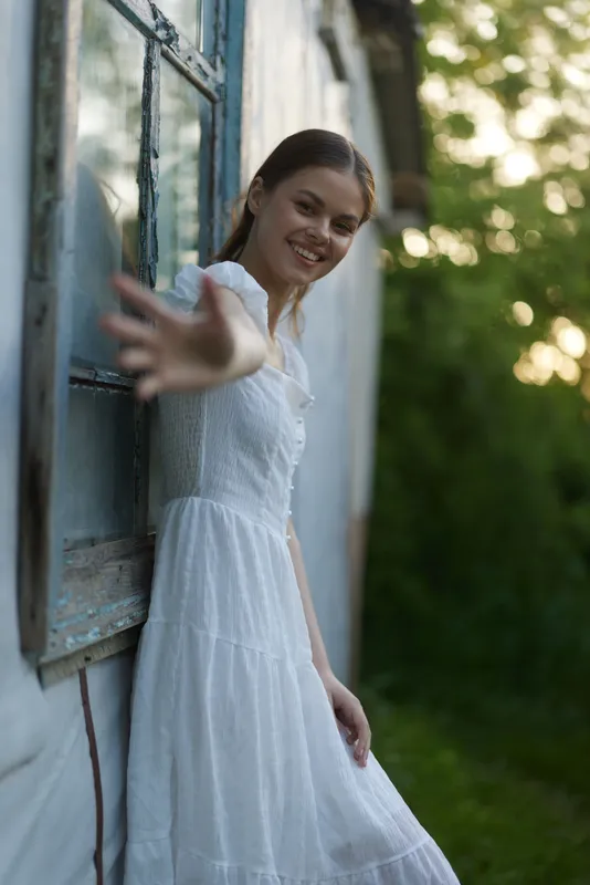Young woman in a white dress smiling and reaching out while standing by a rustic window, surrounded by greenery at sunset