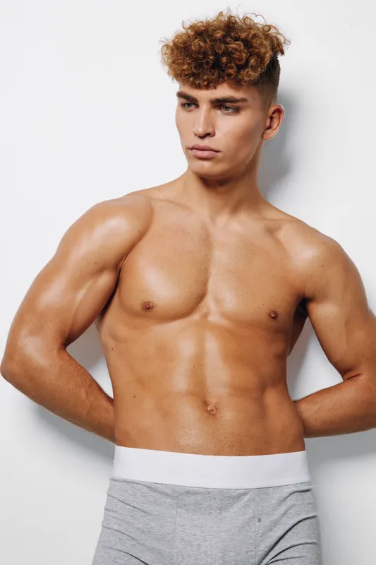 Muscular young man with curly hair posing shirtless against white background showing defined chest and abs with hands behind back in studio lighting.