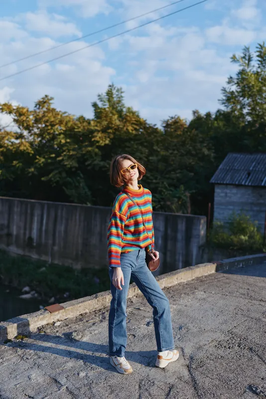 Woman in a colorful striped sweater and sunglasses stands smiling on a rural rooftop, casual outfit with jeans and white sneakers, holding phone for relaxed travel lifestyle vibe.