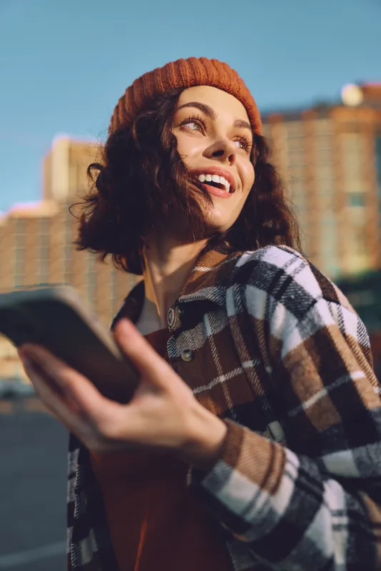 Woman smile with phone in urban sunlight, beanie and plaid jacket, candid moment captured in golden hour glow, authenticity, mindful living and emotional storytelling atmosphere.