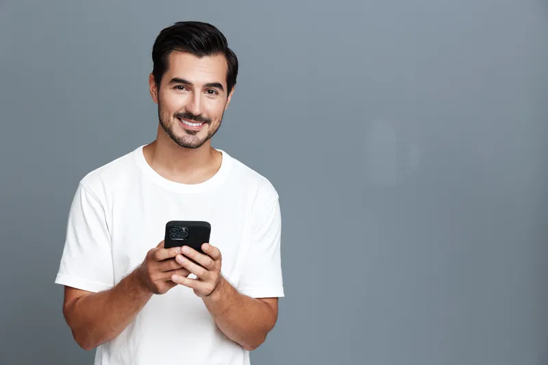 Smiling man in casual white t-shirt looking at camera while holding smartphone in hands, standing against plain solid gray background. People lifestyle concept
