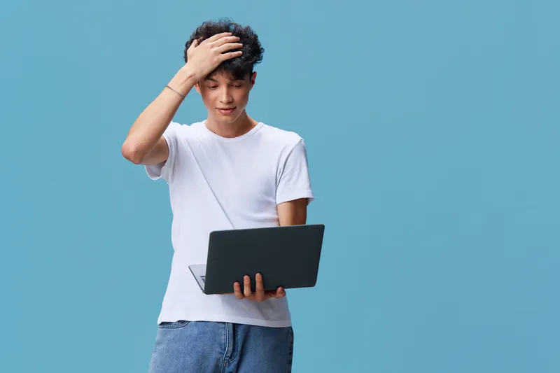 young man holding laptop with thoughtful expression in casual outfit against blue background, technology, communication, modern lifestyle concept