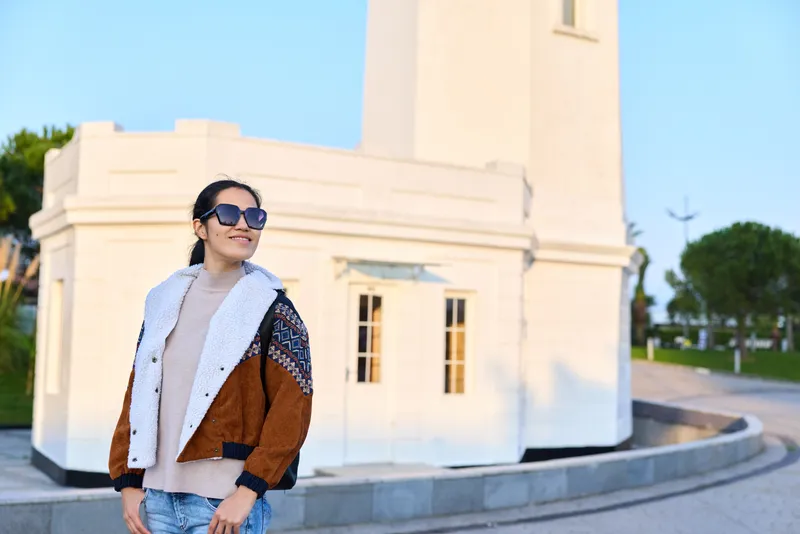 Woman wearing denim and jacket stands outdoors in front of a bright white architectural backdrop, stylish sunglasses and confident pose, capturing modern street fashion and urban scenery