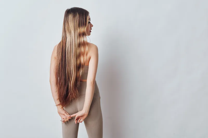 Young woman with long hair standing sideways against a plain background with hands clasped behind back wearing casual outfit and neutral tones.