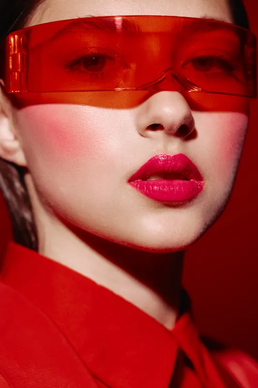 Stylish woman with glossy red lips and bold blush wearing futuristic red tint glasses and red outfit. Close-up beauty portrait against a vivid red background. Concept for fashion, creative, and bold