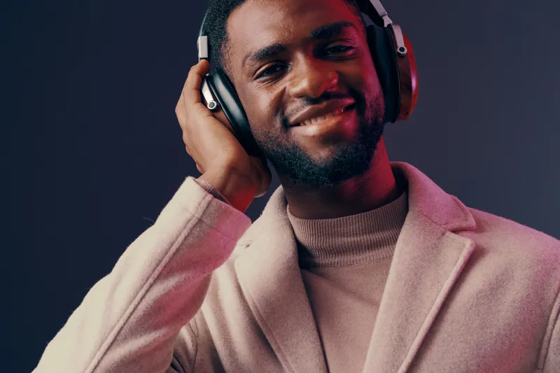 Smiling African man wearing headphones in a stylish coat against a dark gradient background, expressing joy and relaxation in a modern setting
