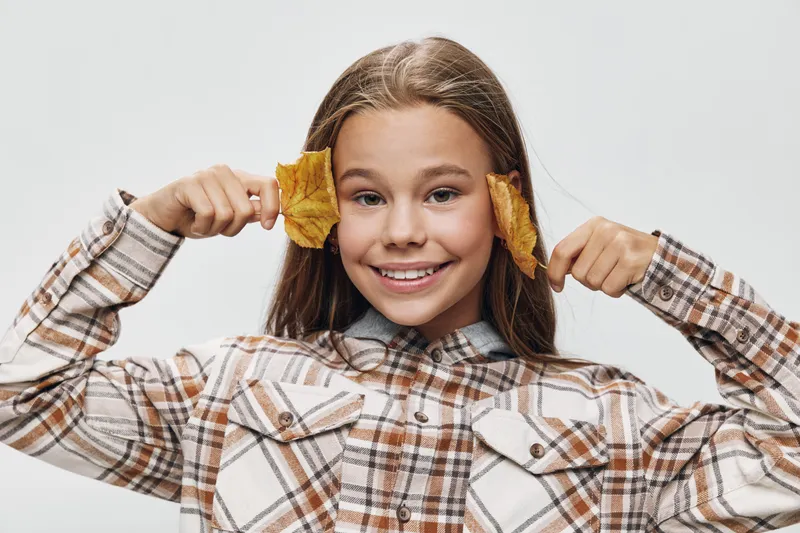 Girl smiling and holding dry autumn leaves near face wearing plaid shirt on white background. Child playing, seasonal nature and happy expression with brown hair.