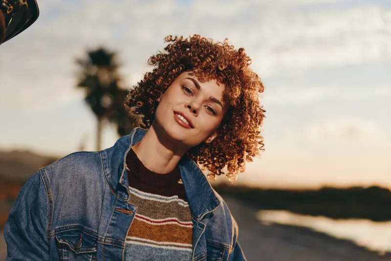 Woman lifestyle outdoors, curly hair in denim jacket, smiling at golden sunset, relaxed stance near coast, warm light, soft breeze, natural colors, calm landscape