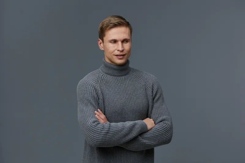 Smiling man wearing gray knitted sweater standing with arms crossed on plain gray background studio portrait. People lifestyle concept, casual style, happiness, confidence, adult male.