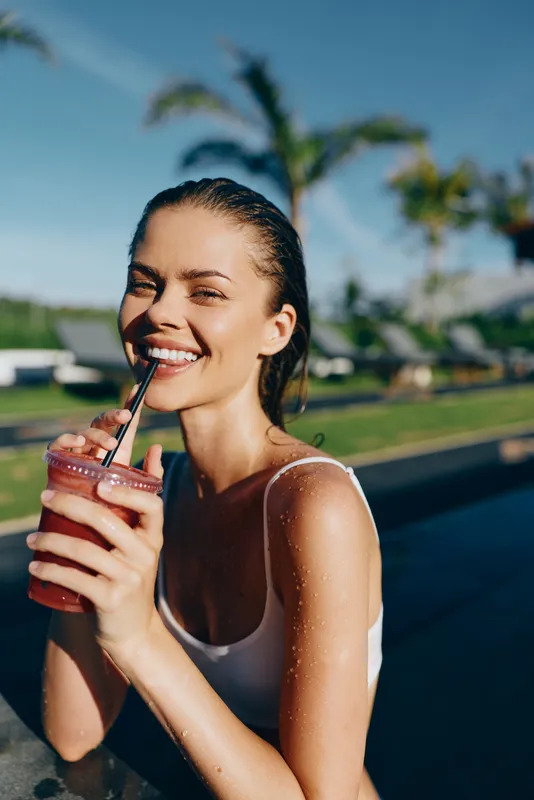 Cheerful woman outdoors, sipping a smoothie by the pool, smiling softly, sunlit moment, casual wellness lifestyle, vibrant colors, refreshing and relaxed. soft breeze around.