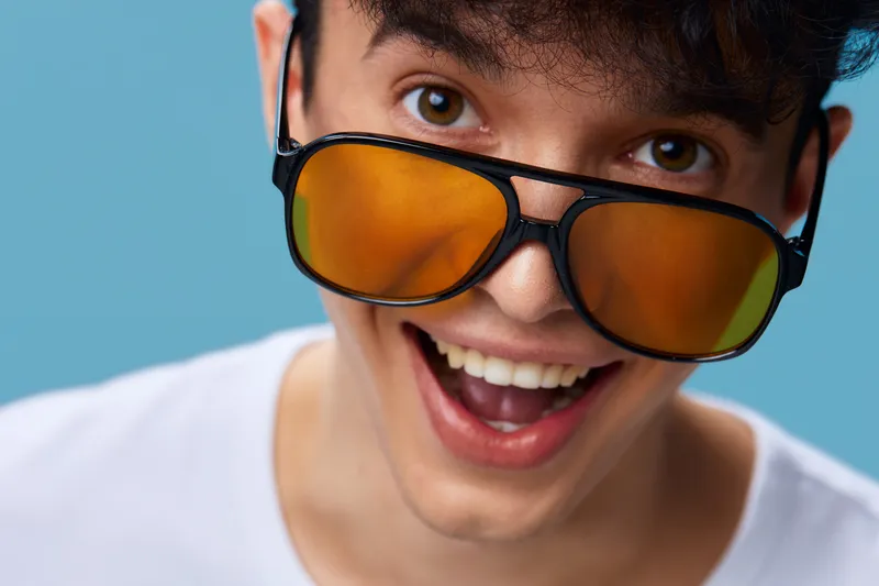 Young man wearing large orange sunglasses smiling joyfully against blue background, expressing happiness and trendy casual style with white t-shirt.