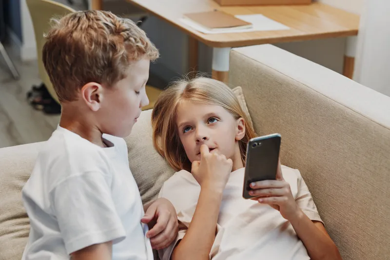 Two children interact on a beige sofa in a cozy room, one holding a smartphone while the other looks attentively. Concept of childhood communication, technology, and family moments.