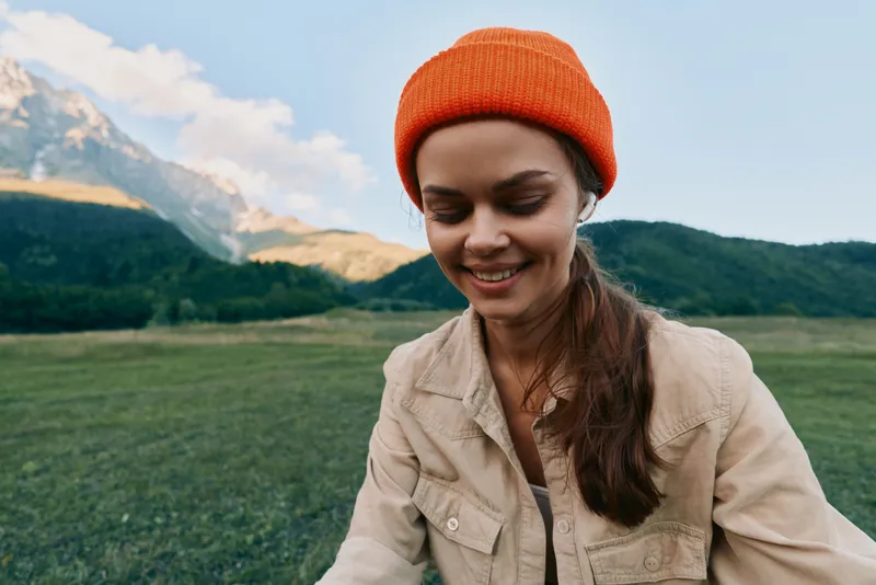 Young woman with long hair sits in a green field wearing an orange beanie and beige jacket, smiling softly as distant mountains rise under a clear blue sky