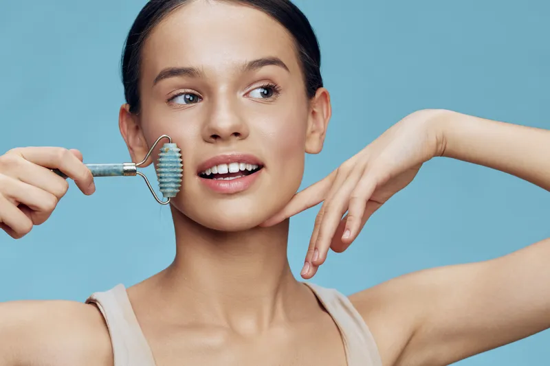 Young woman using a jade roller for skincare, showcasing natural beauty with a joyful expression against a soft blue background, ideal for wellness and beauty themes